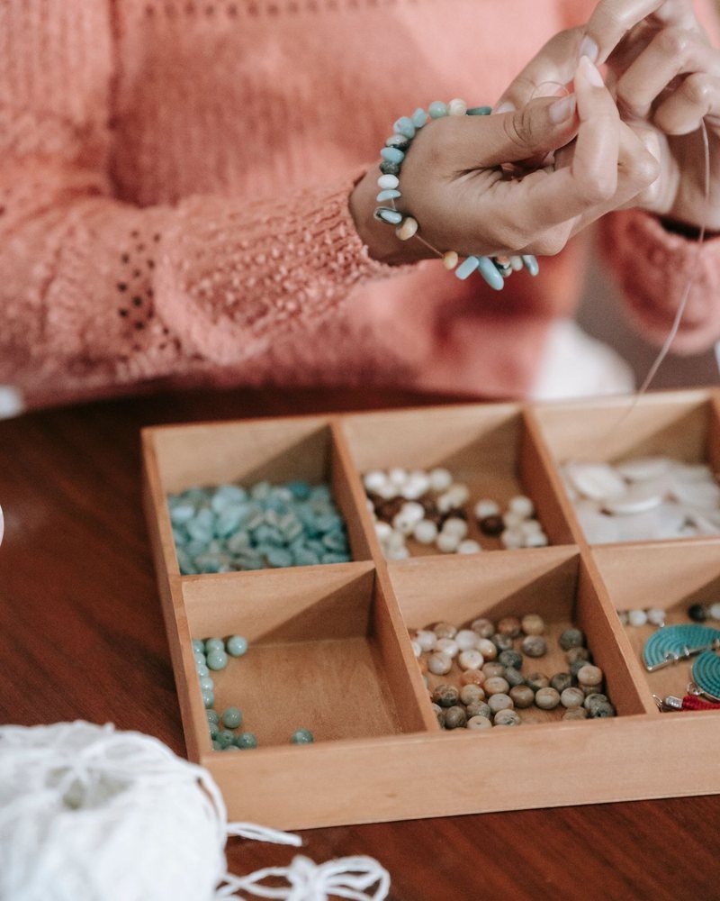 Hands stringing beads above a wooden tray of sorted stones